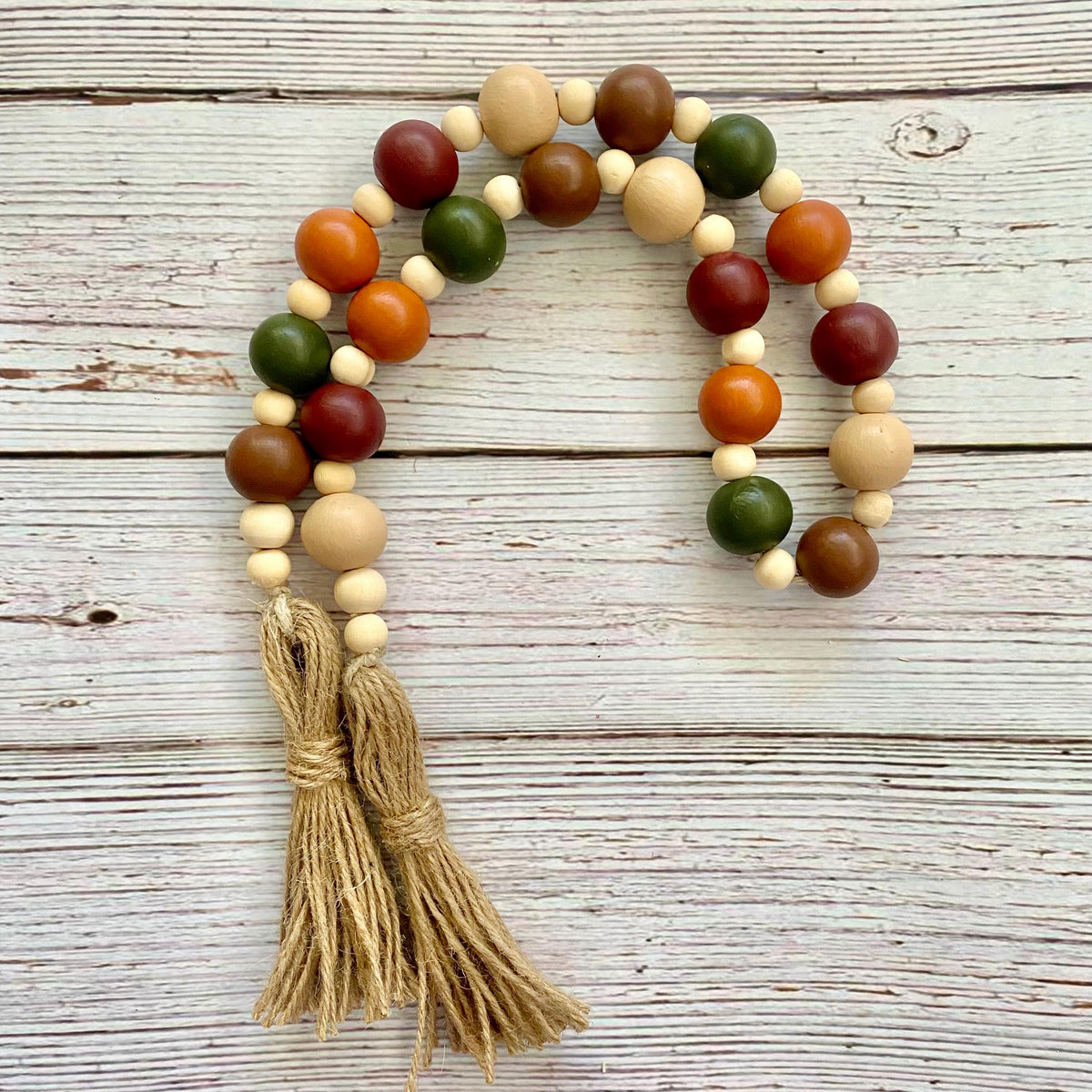 A wooden bead garland with natural beige, brown, orange, green, and dark red beads, finished with two tan cotton tassels, displayed on a rustic white-washed wooden surface.