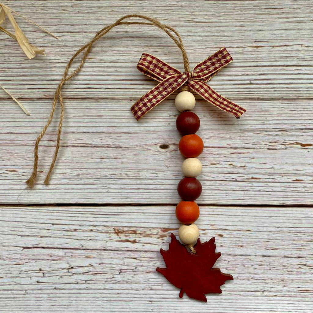 A rustic autumn hanging ornament with a red and white gingham ribbon bow, a string of wooden beads in red, orange, cream, and dark red, and a red maple leaf at the bottom, displayed on a white-washed wood background.