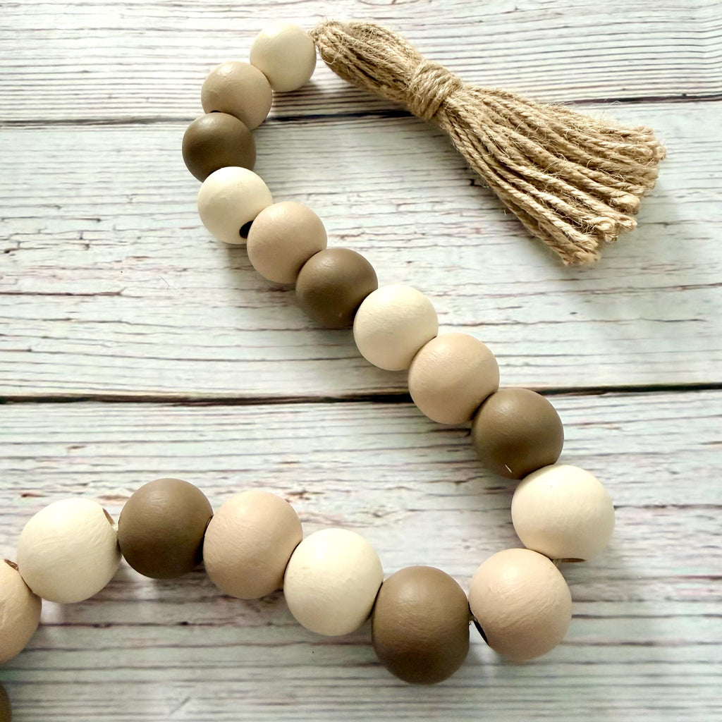 A wooden bead garland with a natural jute tassel, featuring alternating light beige, medium brown, and off-white round wooden beads, laid out on a white-washed wood plank background.