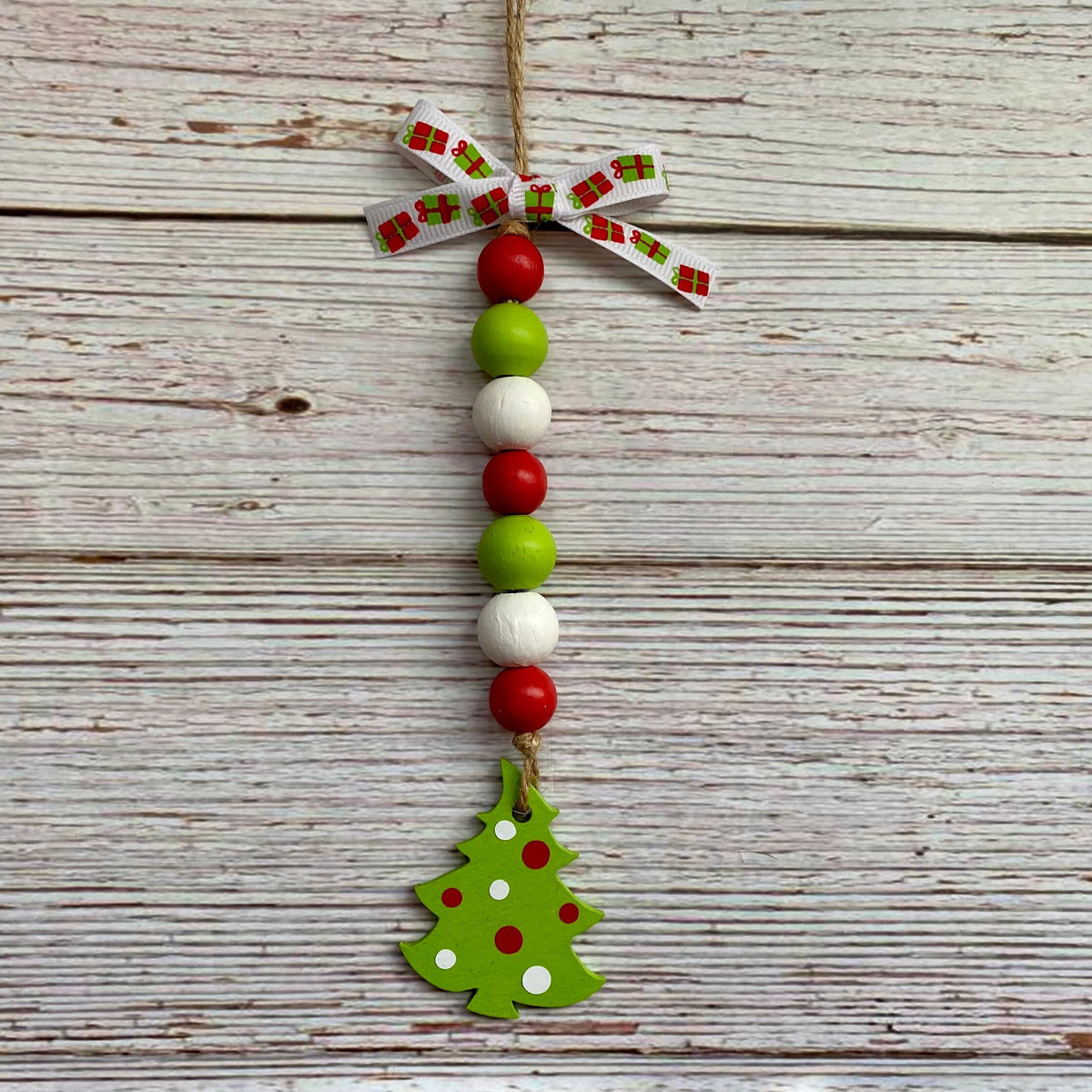A handmade Christmas hanging ornament made of wooden beads in red, green, and white, finished with a green painted Christmas tree charm and a patterned ribbon bow at the top, displayed on a rustic white-washed wood background.