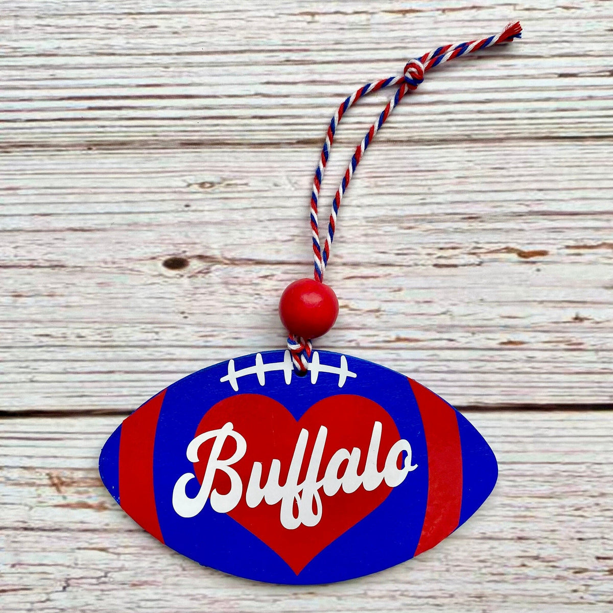 A football-shaped hanging ornament with a red heart, white script text reading 'Buffalo,' and a red, white, and blue cord loop, displayed on a whitewashed wood surface.