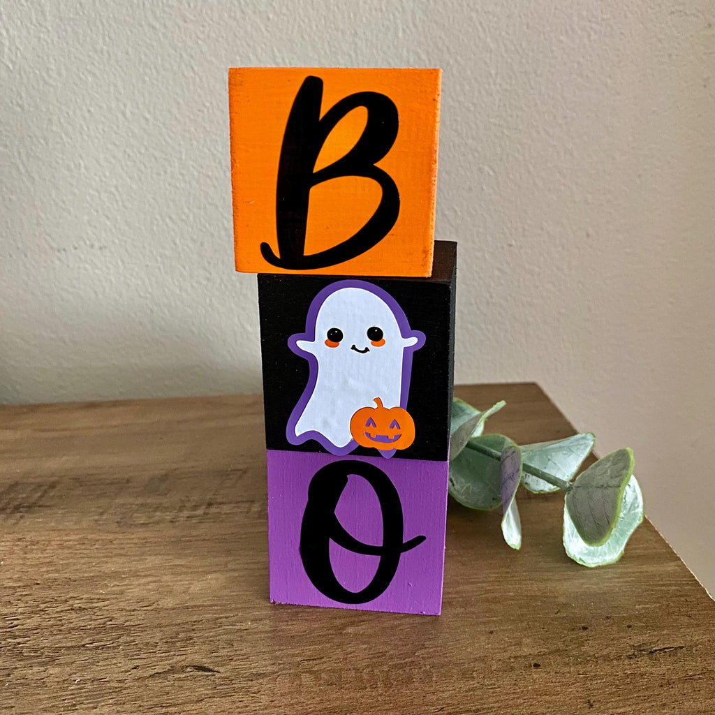Three stacked wooden blocks spelling BOO for Halloween, featuring an orange block with a black letter B, a black block with a cute white ghost holding an orange pumpkin, and a purple block with a black letter O, displayed on a wooden table.