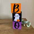 Three stacked wooden blocks spelling BOO for Halloween, featuring an orange block with a black letter B, a black block with a cute white ghost holding an orange pumpkin, and a purple block with a black letter O, displayed on a wooden table.