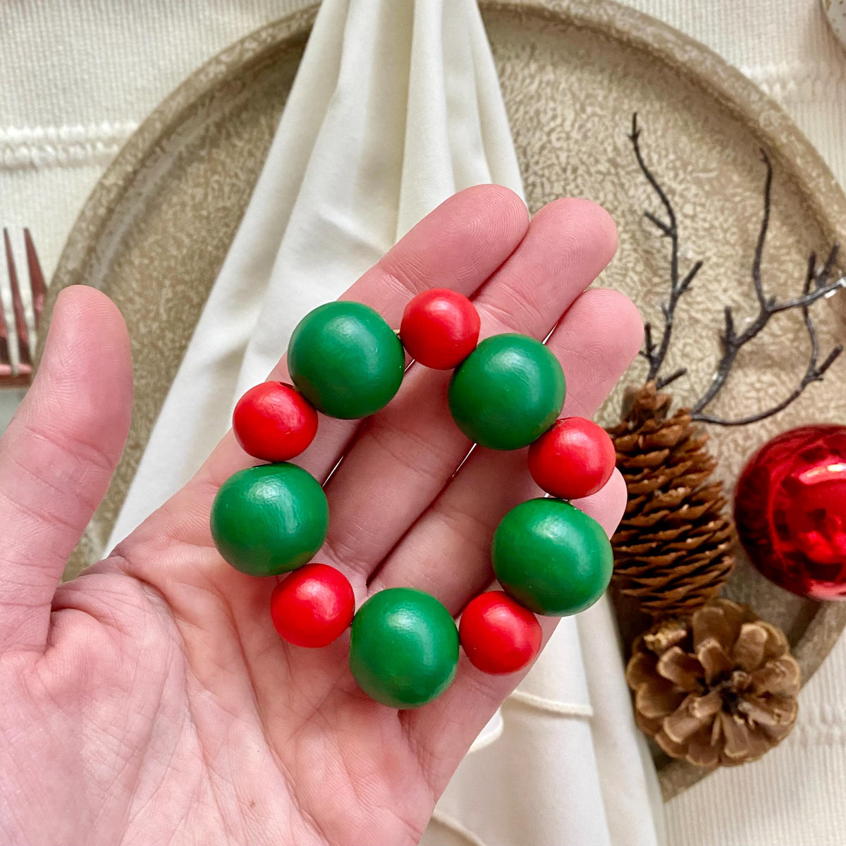 A beaded napkin ring made of alternating green and red round beads, held in a person's hand over a styled holiday table setting with a napkin, pinecones, and a red ornament.