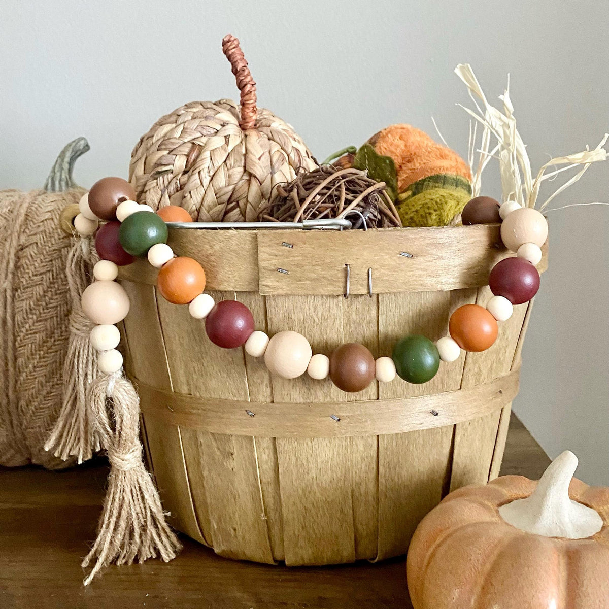 A wooden bead garland with autumn colors hangs around a rustic wooden basket filled with fall decor, including woven pumpkins and dried grass, on a tabletop.