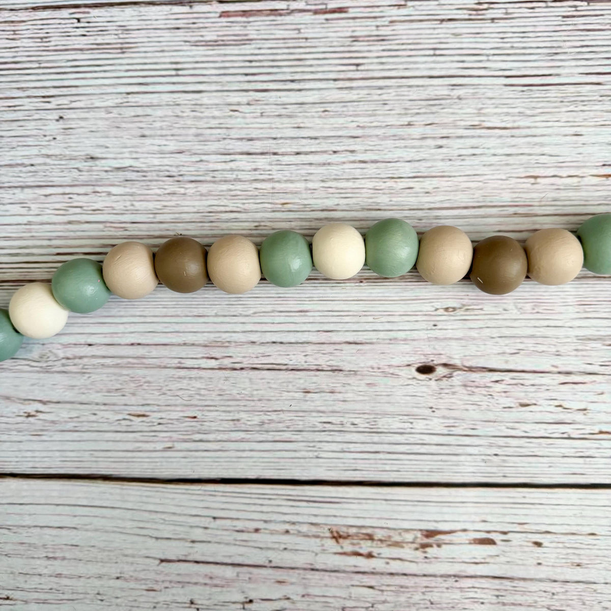 A string of matte wooden beads in muted sage green, cream, tan, and brown, laid horizontally on a distressed white wood plank surface.