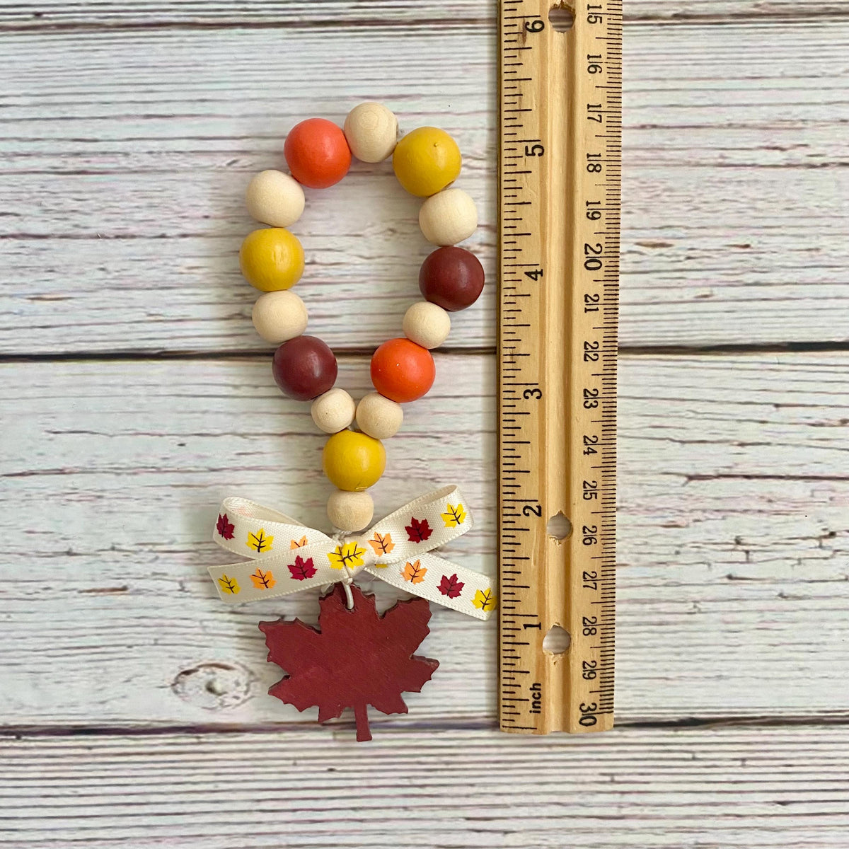 A beaded wood bead loop made of alternating yellow, orange, cream, and dark red wooden beads, with a cream ribbon printed with autumn leaves and a red maple leaf charm, shown next to a wooden ruler on a whitewashed wood background.