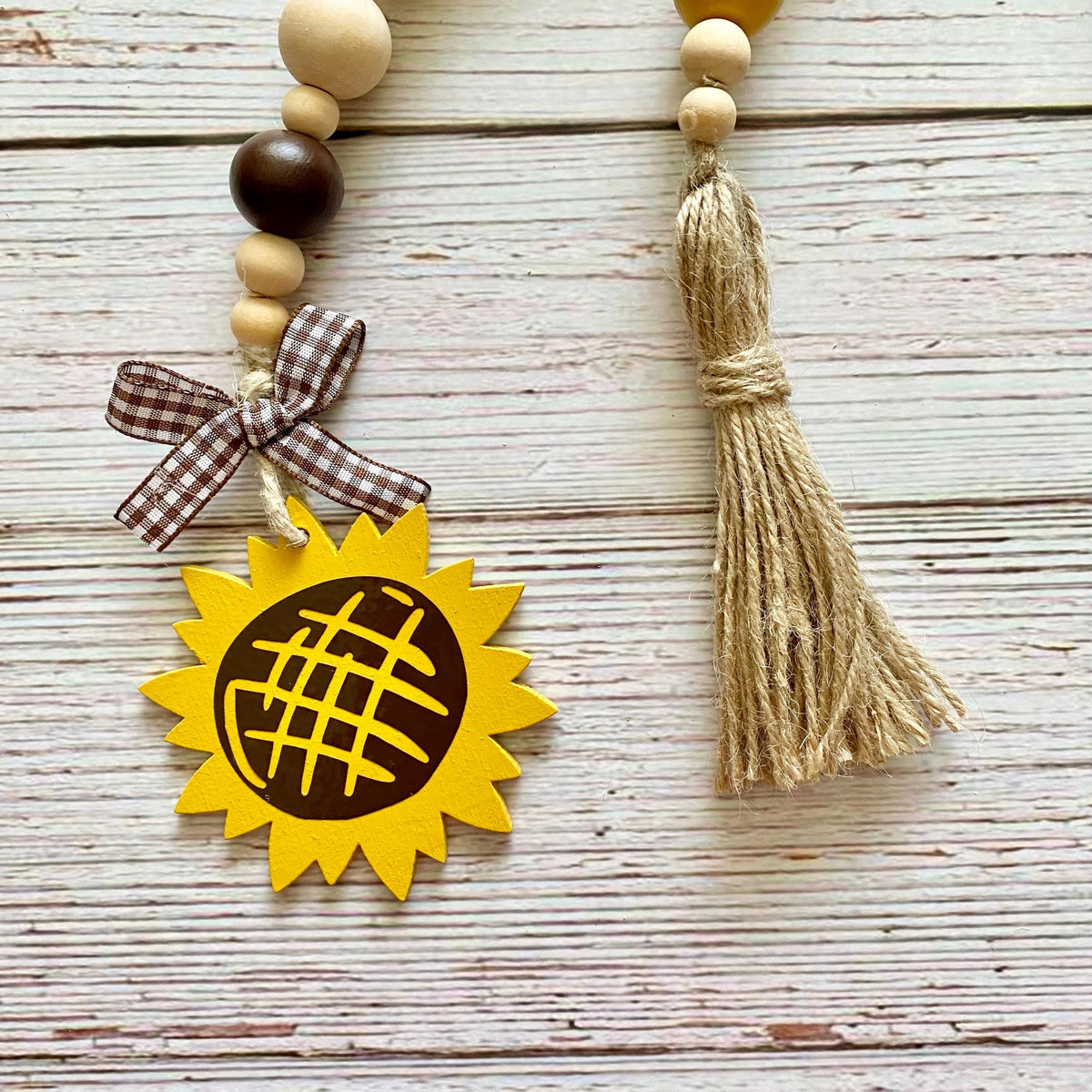 A rustic wooden bead garland with a sunflower charm, a brown gingham ribbon bow, and a natural jute tassel, displayed on a whitewashed wooden plank background.