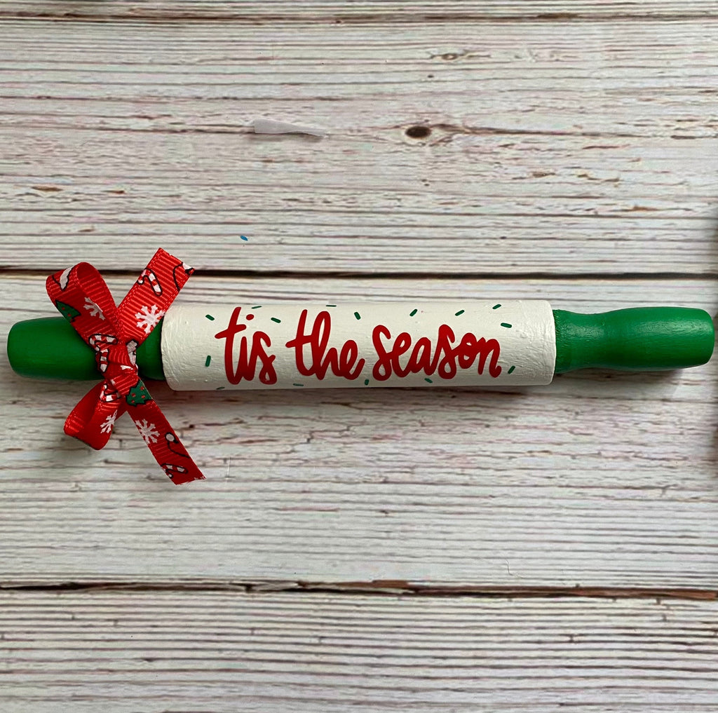 A Christmas-themed wooden rolling pin with green handles, a white center wrapped in a red ribbon, and 'Tis the season' written in red script. The rolling pin is placed on a white-washed wooden plank background next to a red, green, and white wooden bead garland.