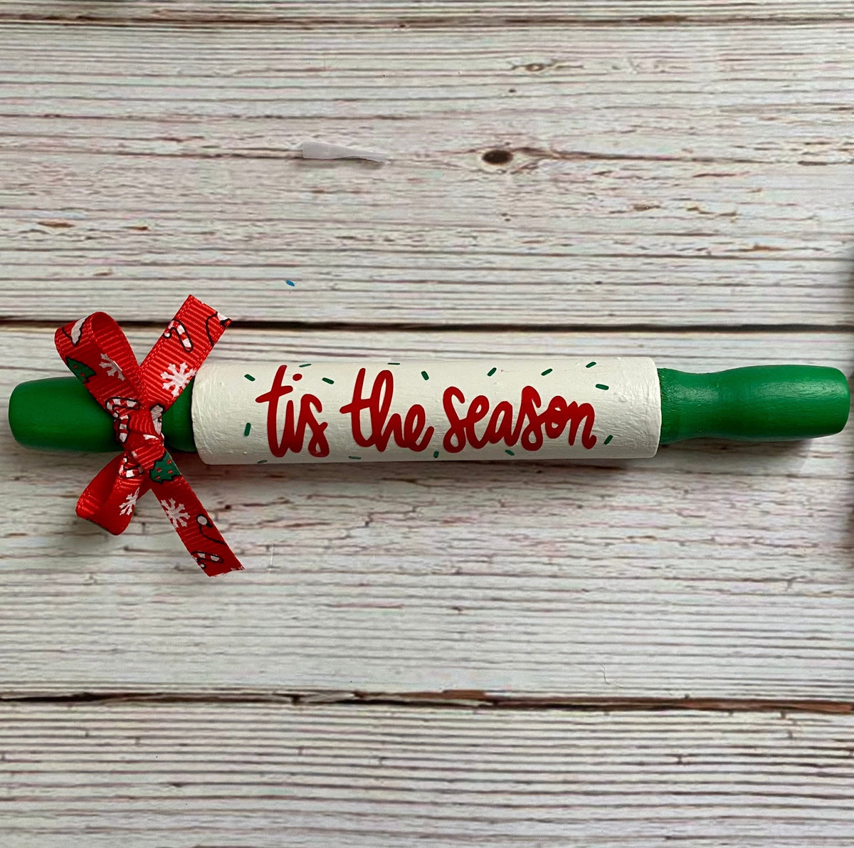 A Christmas-themed wooden rolling pin with green handles, a white center wrapped in a red ribbon, and 'Tis the season' written in red script. The rolling pin is placed on a white-washed wooden plank background next to a red, green, and white wooden bead garland.