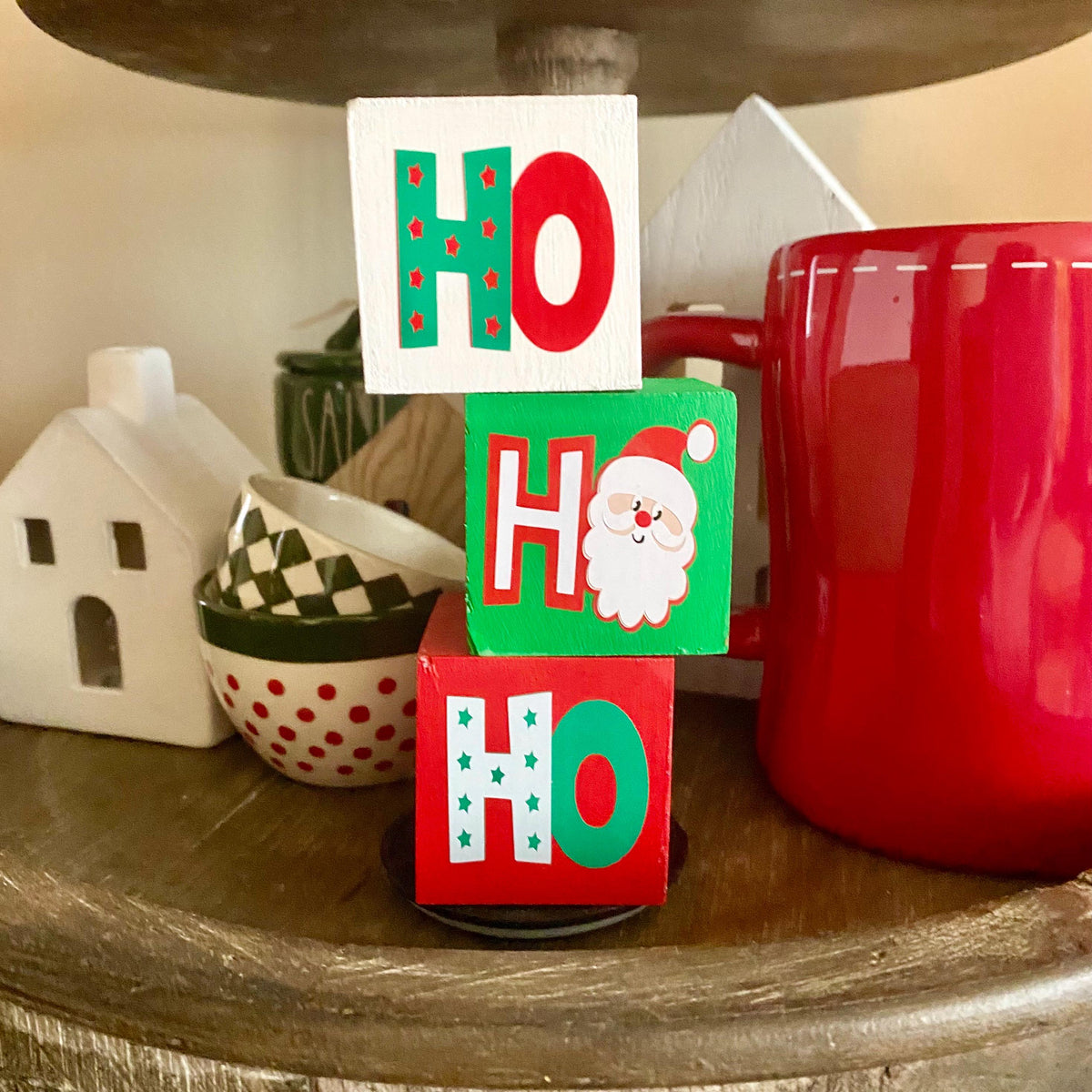 Three stacked wooden decorative blocks spelling HO HO HO in a holiday setting on a wooden shelf, with a red mug and small white house decor in the background.