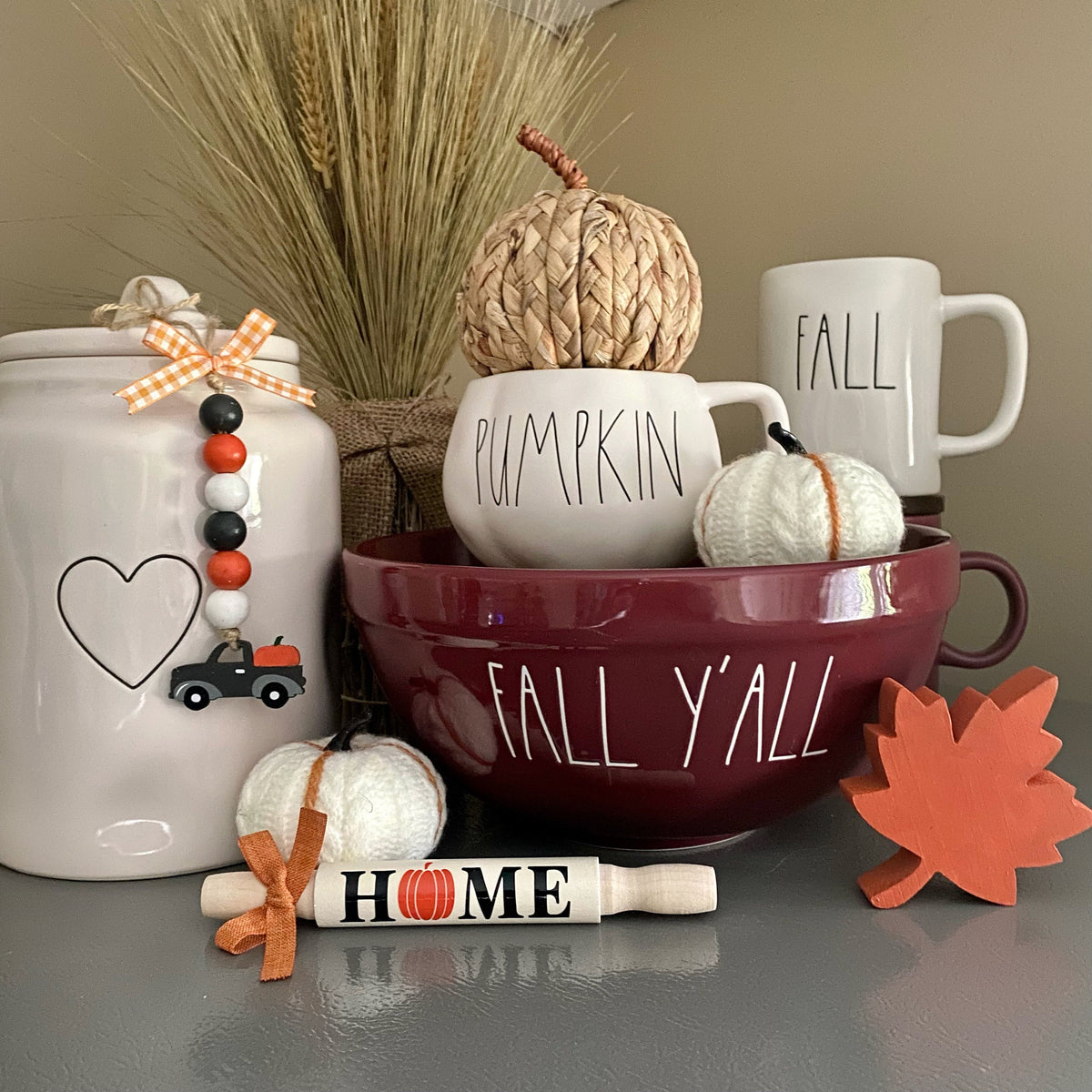 A styled autumn tabletop arrangement featuring a white ceramic jar with a beaded bow and heart design, a large burgundy bowl with white 'FALL Y'ALL' lettering, stacked white mugs reading 'PUMPKIN' and 'FALL', a woven pumpkin, a small white pumpkin, a wooden 'HOME' rolling pin ornament, and an orange leaf decoration.