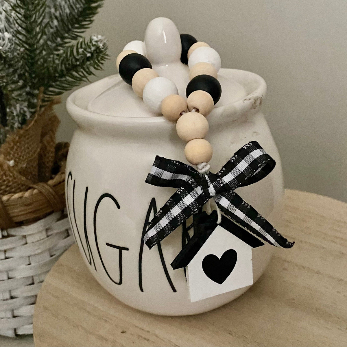 A cream-colored ceramic sugar bowl decorated with a black and white gingham ribbon bow, a small white tag with a black heart, and a beaded garland draped over the lid. The bowl is placed on a light wooden table next to a potted artificial pine tree.