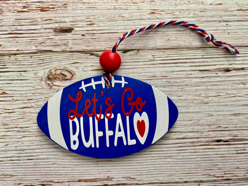A football-shaped decorative ornament with a blue and white painted finish, white laces, red script text reading 'Let's Go BUFFALO,' a small red heart, and a red-and-white hanging cord, displayed on a rustic wood surface.