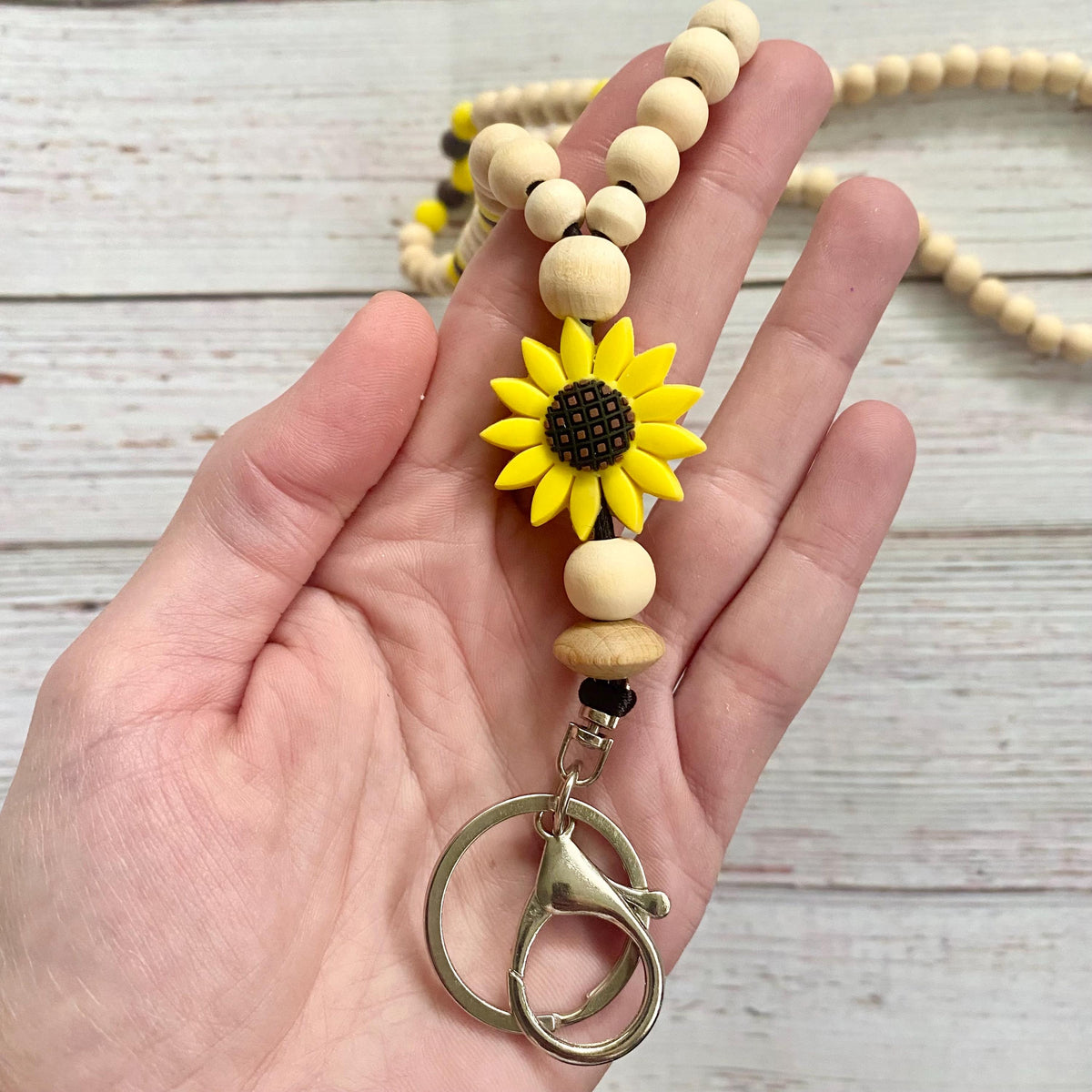 A hand holding a beaded keychain with a yellow sunflower charm, light wood beads, and silver key rings, set against a light wood plank background.