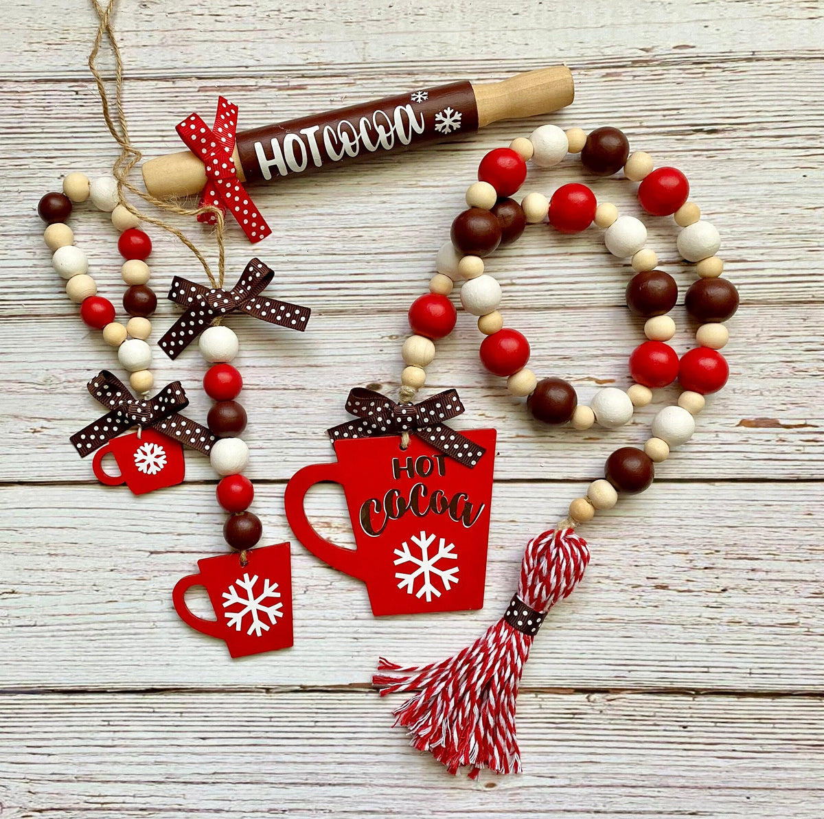A rustic holiday garland set featuring red and white beaded garlands with a wooden rolling pin ornament, red mug and hot cocoa cutouts, brown polka dot bows, and a red-and-white tassel, arranged on a whitewashed wooden surface.