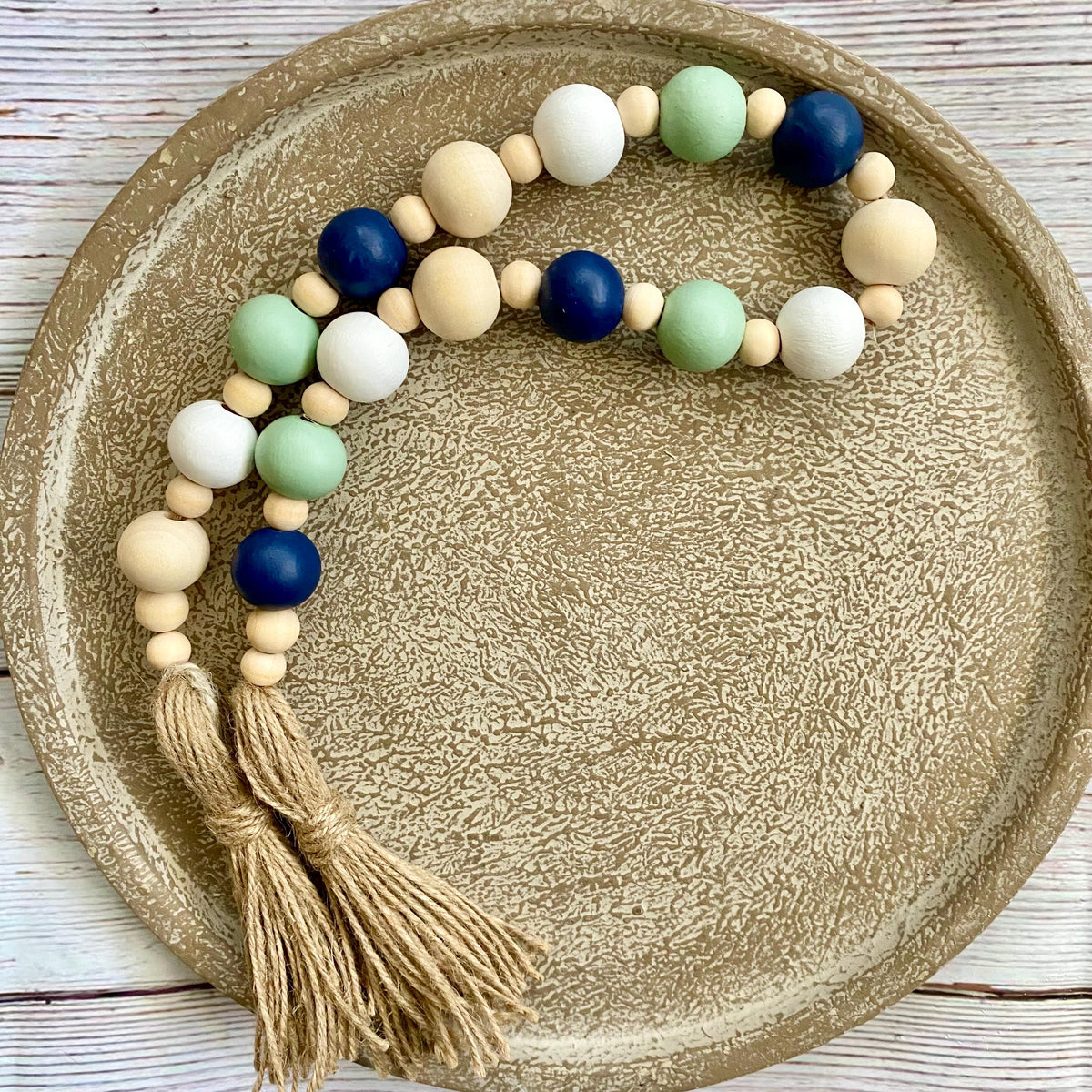 A beaded garland with natural wood beads, navy blue beads, mint green beads, and white beads, finished with two tan rope tassels, displayed on a textured beige ceramic plate.