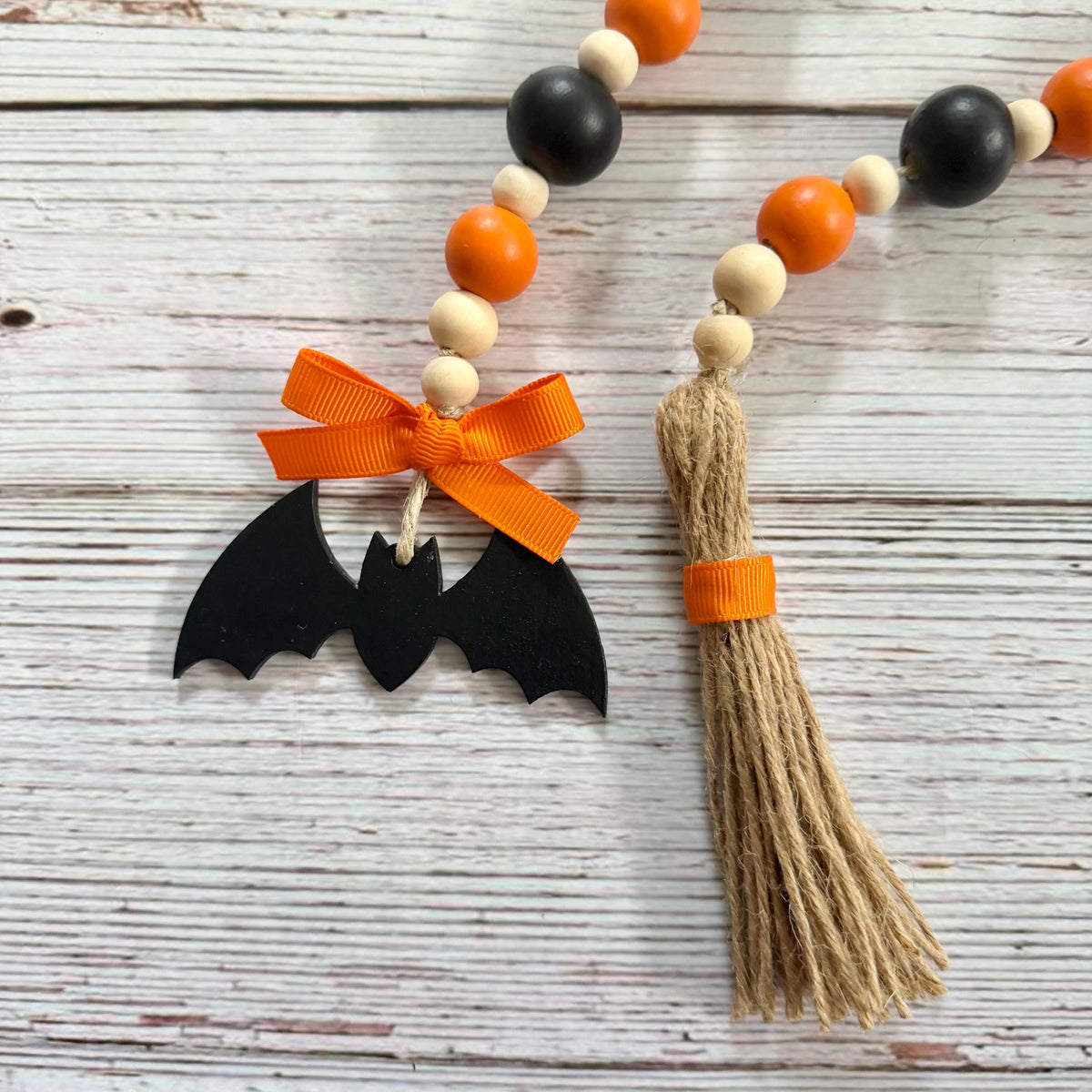 A Halloween-themed beaded garland laid on a whitewashed wooden surface, featuring orange and cream wooden beads, black beads, a black bat silhouette charm tied with an orange ribbon, and a tan jute tassel with an orange band.