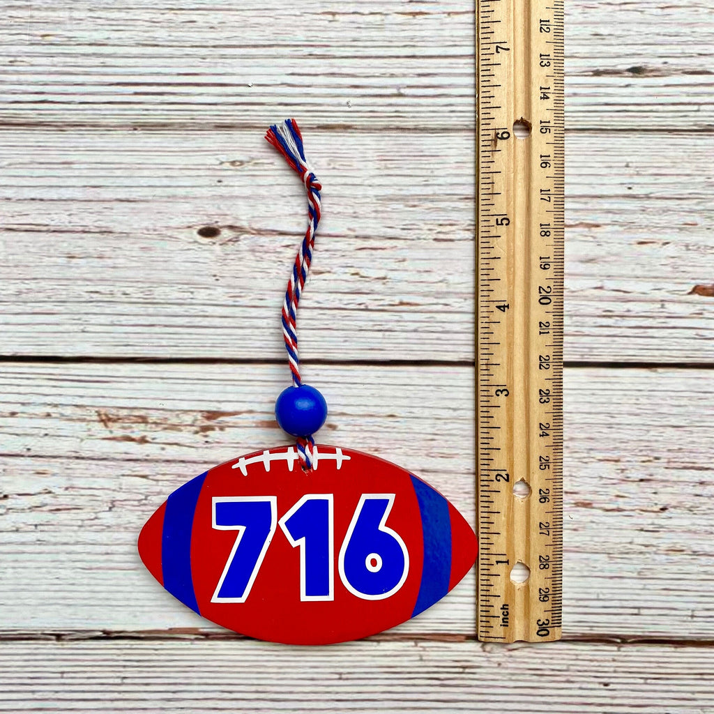 A red and blue football-shaped ornament with the number 716 in white and blue, hanging next to a wooden ruler on a whitewashed wood plank background.