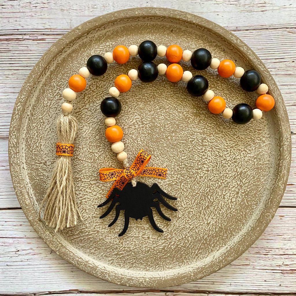 A beaded garland with black and orange wooden beads, a beige tassel, and a black spider ornament, displayed on a round textured gold tray over a white wood background.