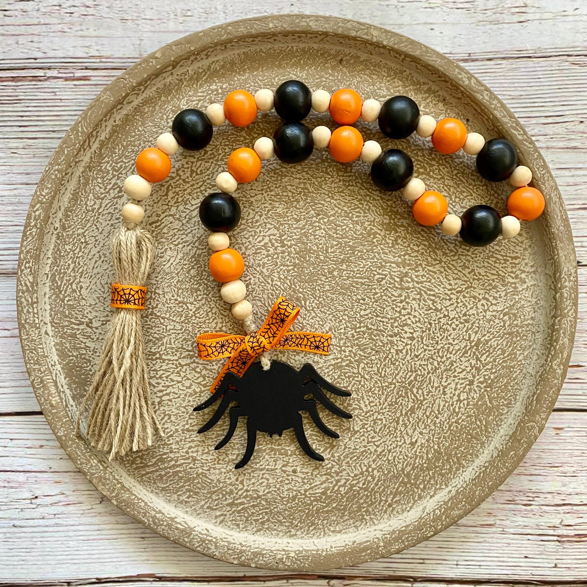 A beaded garland with black and orange wooden beads, a beige tassel, and a black spider ornament, displayed on a round textured gold tray over a white wood background.