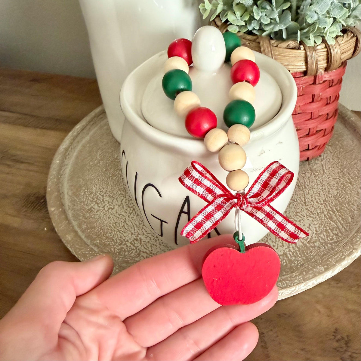 A hand holds a red wooden apple-shaped ornament dangling from a beaded garland with red, green, and natural wood beads, tied with a red-and-white gingham ribbon bow. The scene is styled with a white ceramic canister and a potted succulent in the background.